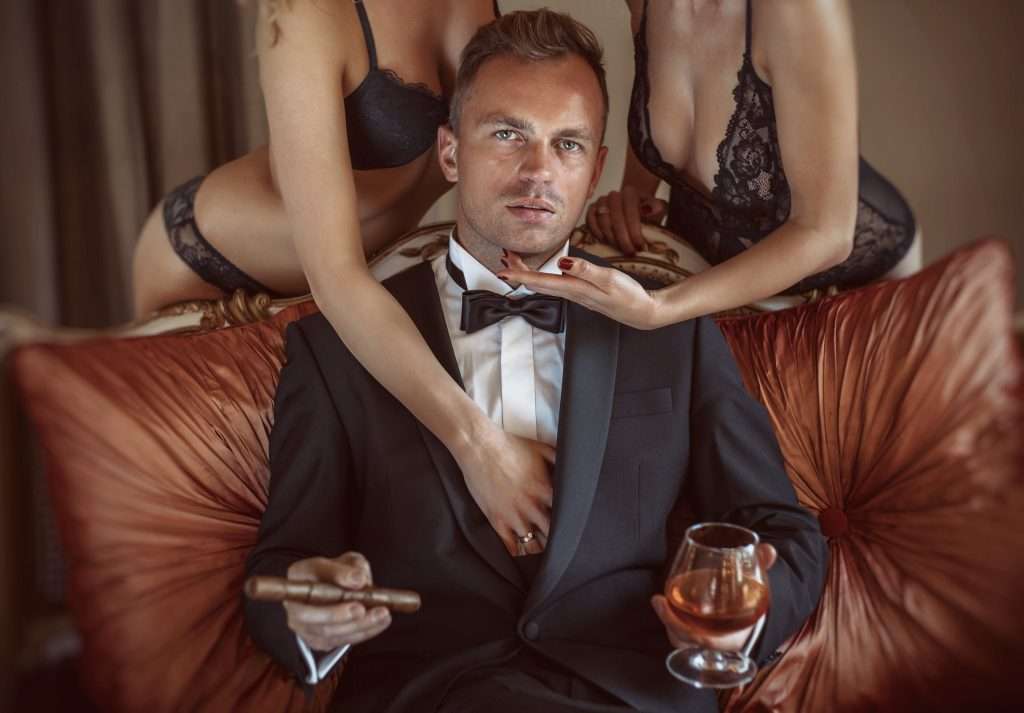 Generous Brisbane VIP client enjoying luxury escort experience with two stunning VIP Escorts, holding a glass of whisky and a cigar